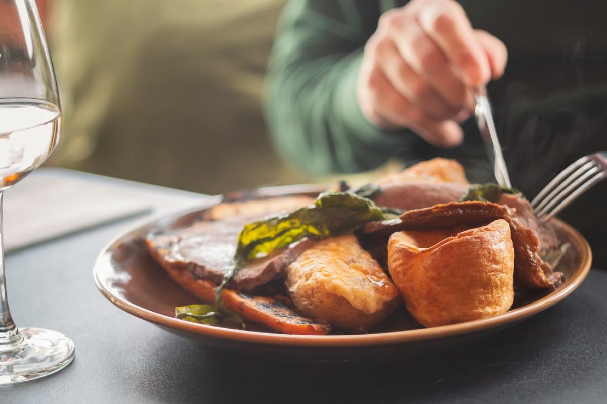 Shallow focus on  yorkshire pudding as part of a meal of roast beef and vegetables. A mans hands can be seen using a knife and fork. A wine glass is on the table.